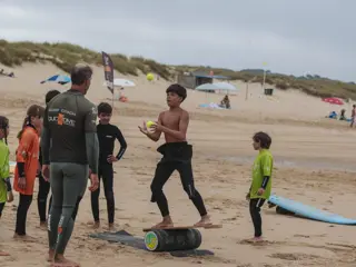 Um treinador da Duckdive com um grupo de crianças na areia; uma delas equilibra-se numa prancha sobre um rolo a apanhar uma bola
