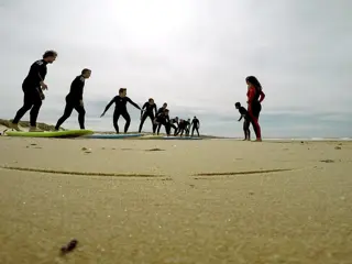 Uma fila de alunos a treinar o take off em cima das pranchas na areia, com o treinador a acompanhar