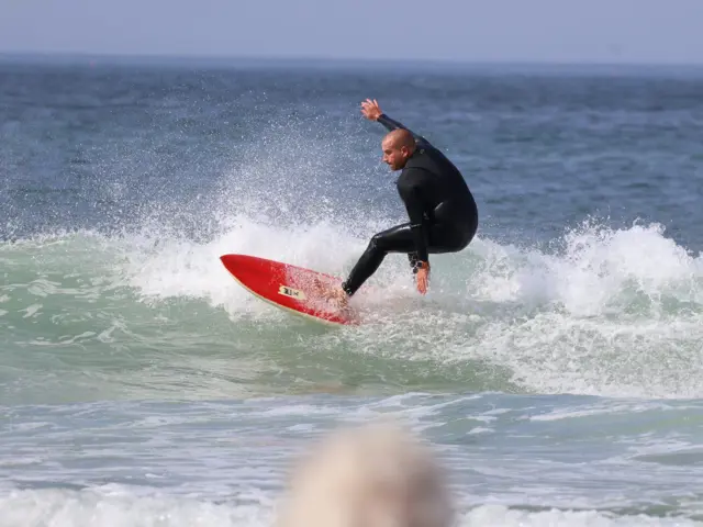 Um surfista de fato preto a cortar a parede de uma onda numa prancha vermelha