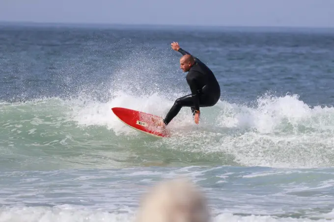 Um surfista de fato preto a cortar a parede de uma onda numa prancha vermelha