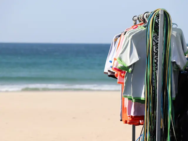 Licras e fatos pendurados num bengaleiro na praia, com o mar em fundo